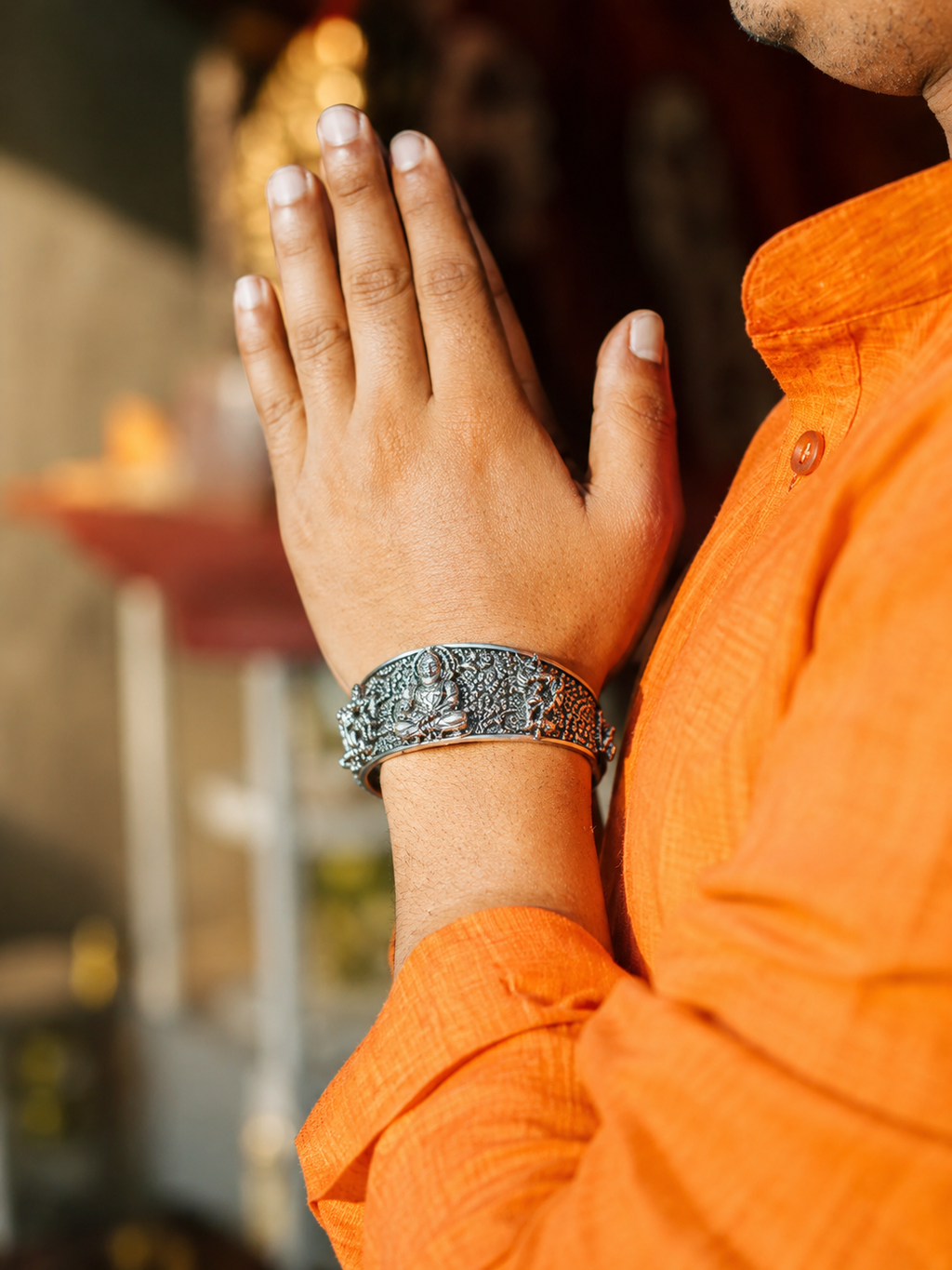 Person wearing an orange outfit with a silver bracelet, hands clasped together in a gesture of prayer or respect.